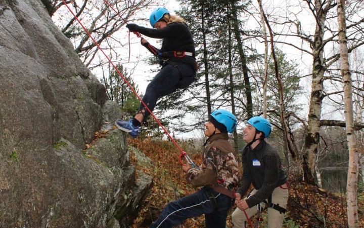 Three people wearing safety gear participate in rock climbing during the family seminar of an outward bound intercept course.